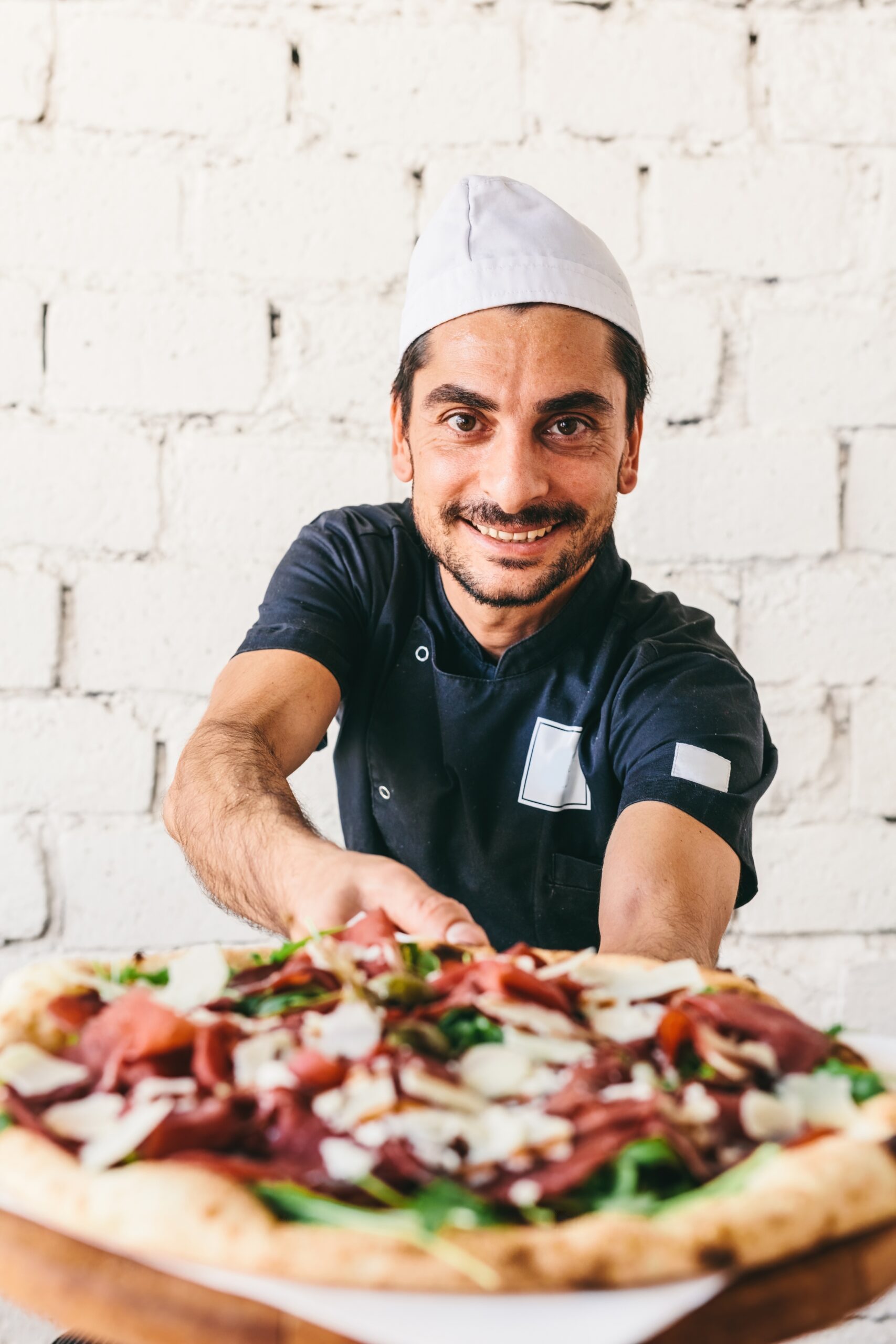 Smiling,Italian,Chef,Or,Pizzaiolo,Offering,Pizza,In,Restaurant